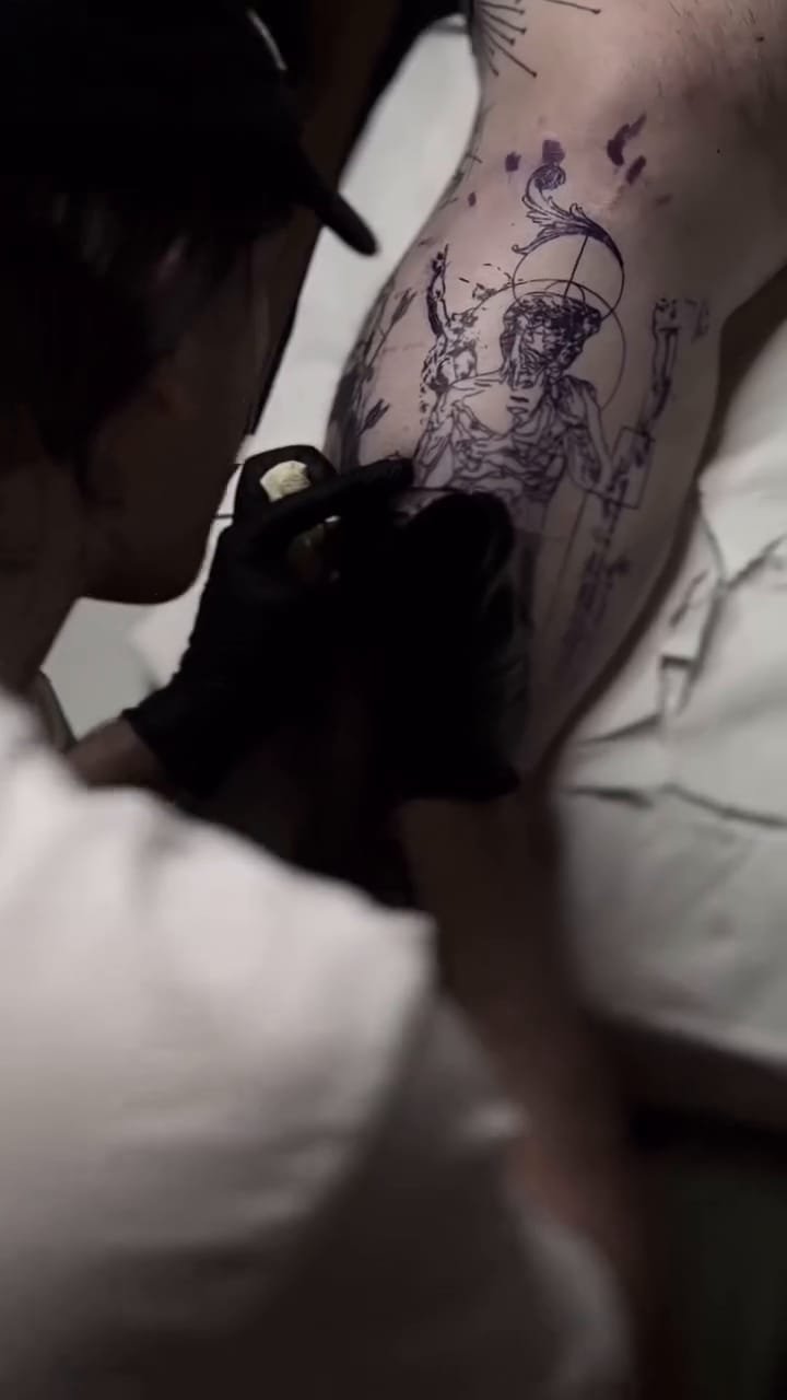 Tomás, con gorra y guantes negros, tatúa la pierna sobre el stencil violeta del David; después, primeros planos de la aguja y el resultado: las piernas de la estatua sobre su pedestal y columnas de texto manuscrito.
