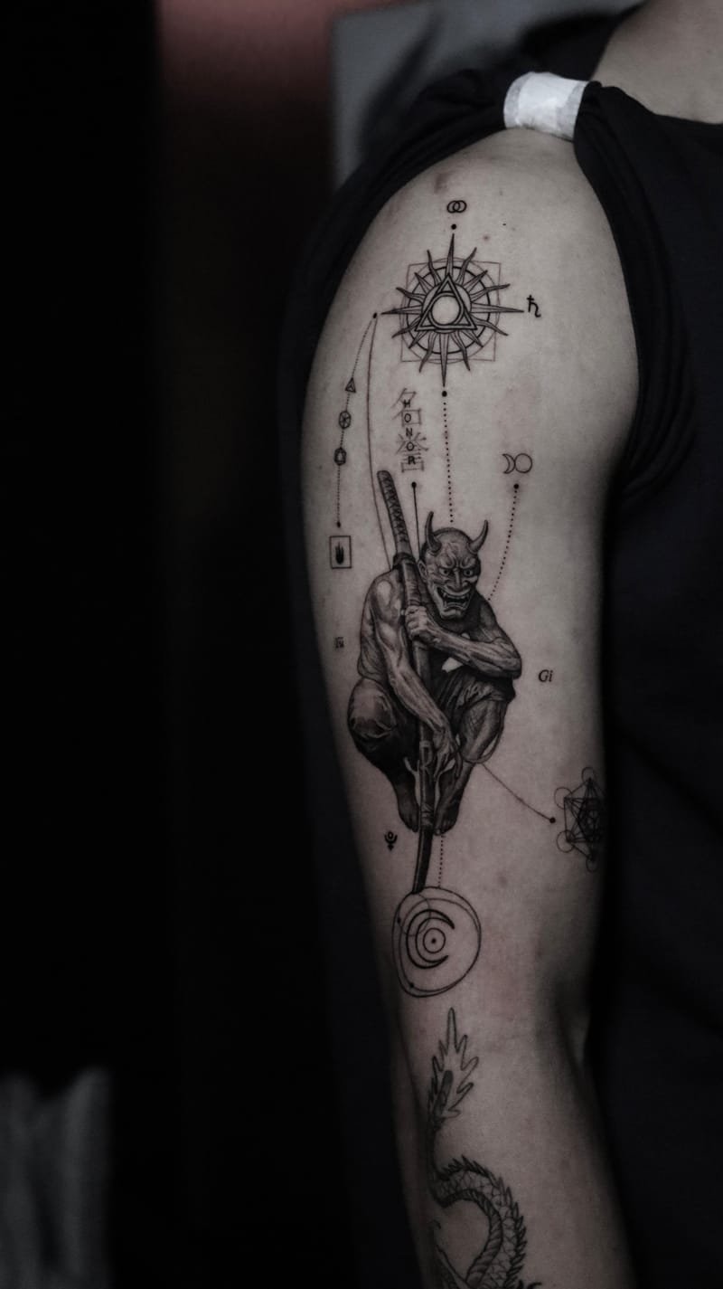 Brazo tatuado con una figura agazapada que lleva una máscara hannya de cuernos y sostiene una katana apoyada en el suelo; por encima, una rosa de los vientos con un triángulo inscrito y, alrededor, símbolos planetarios unidos por líneas punteadas.